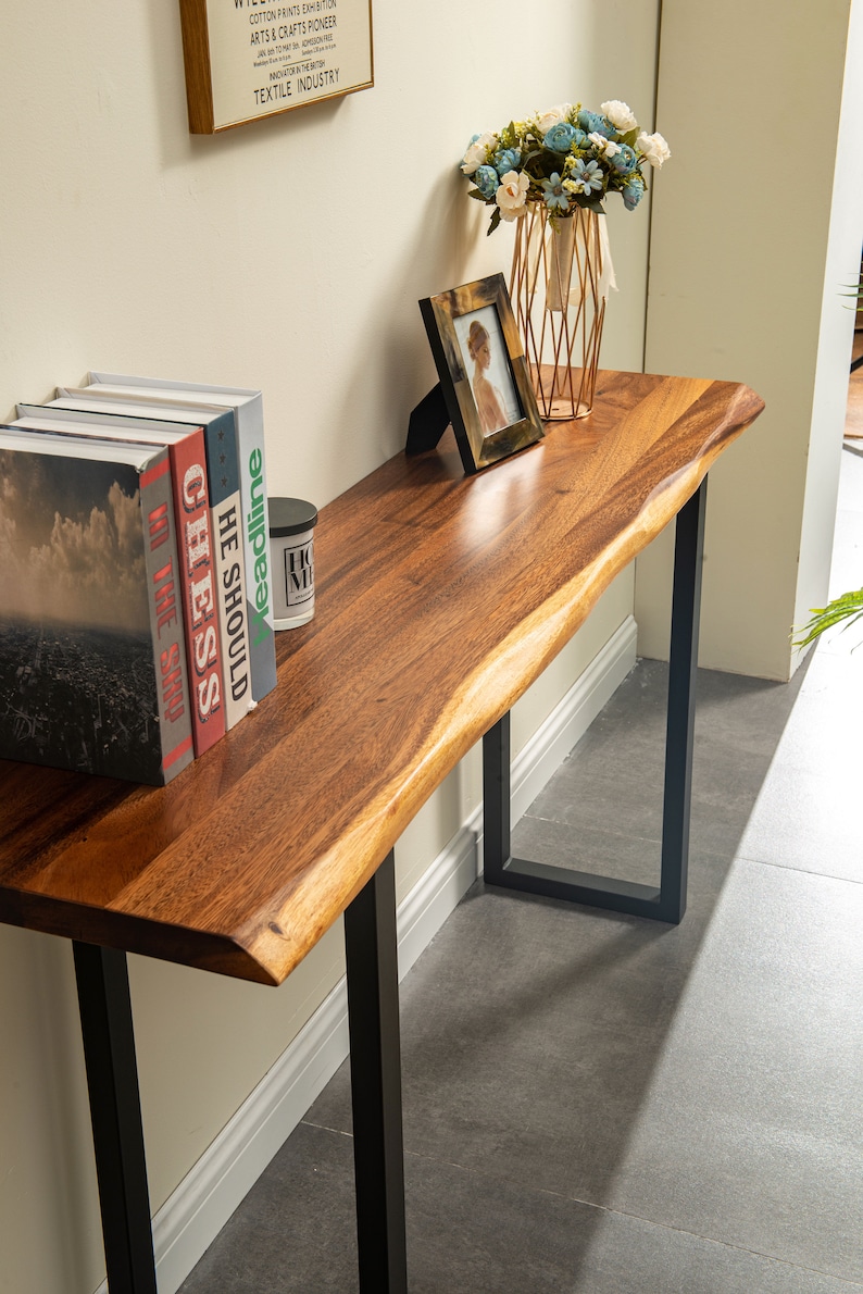 May include: A long, wooden console table with a live edge top and black metal legs. The table is decorated with a vase of flowers, a framed photo, and a stack of books with titles like "Chess" and "He Should".