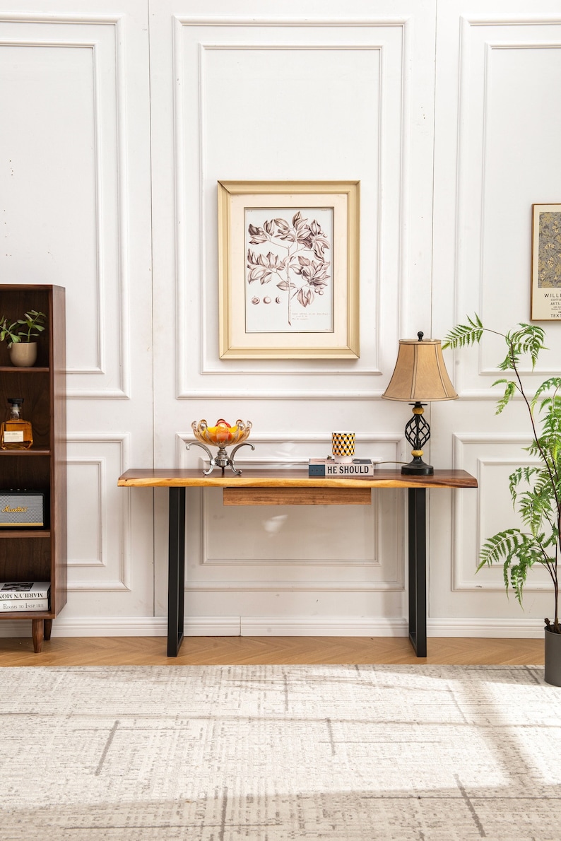 May include: A wooden console table with black metal legs. The table has a drawer and is decorated with a bowl of fruit, a book, and a lamp.