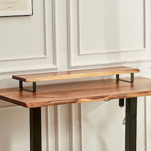 May include: A wooden desk riser with black metal brackets. The riser is made of natural wood with a live edge and is sitting on top of a wooden desk with black metal legs.