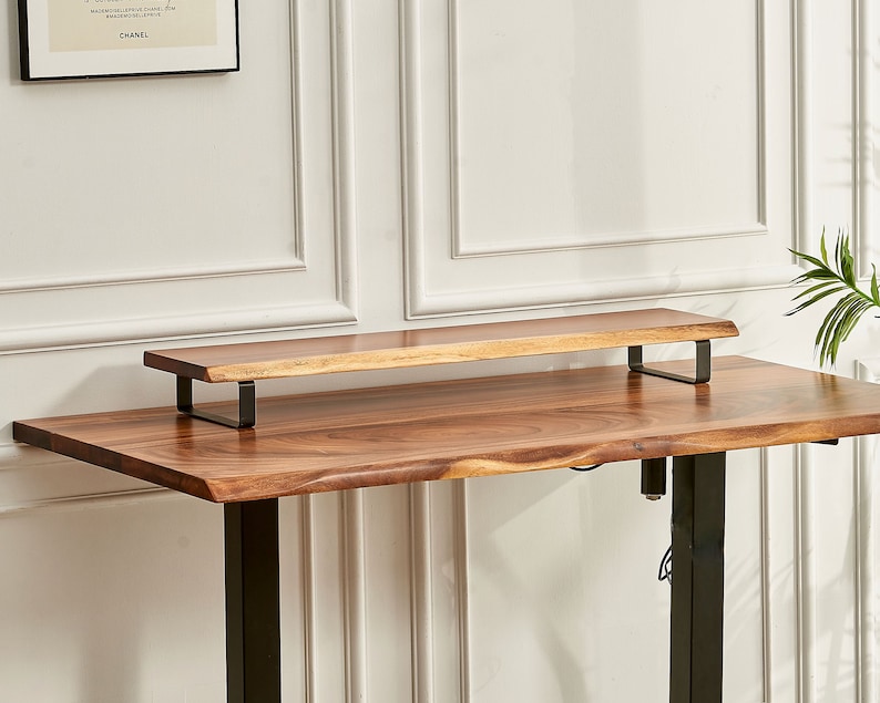 May include: A wooden desk riser with black metal brackets. The riser is made of natural wood with a live edge and is sitting on top of a wooden desk with black metal legs.