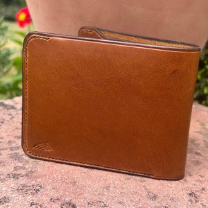 May include: A tan leather wallet with a bi-fold design. The wallet is a warm, medium tan colour with a smooth texture. The edges are neatly stitched, and a small embossed design is visible on the lower right corner.