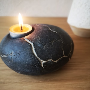 Op de afbeelding: Een zwarte, steenachtige kandelaar met een brandend waxinelichtje. De houder heeft een afgeronde vorm met een gebarsten, getextureerd oppervlak en een witte accent. De vlam van de kaars is heldergeel. Op de achtergrond staat een witte vaas.