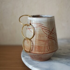 Könnte beinhalten: Eine handgefertigte Keramik-Tasse mit einem einzigartigen Design. Die Tasse hat einen beigefarbenen Grundton mit rotbraunen linearen Mustern und einem weißen Rand. Ein goldfarbener Griff aus drei verbundenen Ringen sorgt für einen modernen Touch. Die Tasse steht auf einem Marmortablett.