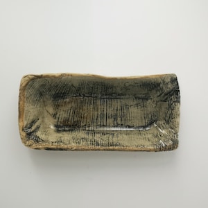 Può includere: Piatto rettangolare in ceramica con superficie strutturata. Il piatto ha una base beige chiaro con segni grigio scuro e neri. I bordi sono leggermente ruvidi, conferendogli un aspetto rustico. Il piatto è vuoto.