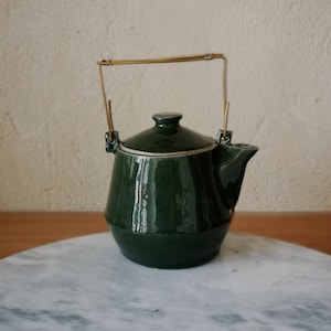 Könnte beinhalten: Eine dunkelgrüne Keramik-Teekanne mit einem goldfarbenen Griff. Die Teekanne hat einen Deckel und einen Ausguss. Der Griff ist mit kleinen Metallbügeln am Körper befestigt. Die Teekanne steht auf einer Marmoroberfläche.