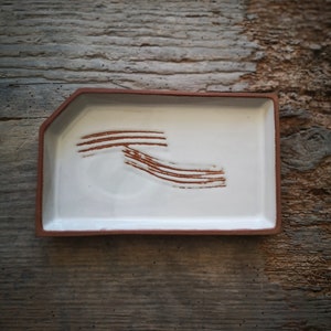 May include: A rectangular ceramic tray with a white interior and a reddish-brown rim. The tray features a series of brown, curved lines on the interior. The tray is set against a rustic, wooden background, showcasing a handcrafted aesthetic.