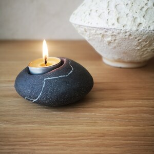 Puede incluir: Una vela de té encendida se encuentra en un portavelas de color gris oscuro, similar a una piedra, con un diseño craquelado blanco. Un jarrón blanco texturizado está en el fondo. La llama de la vela brilla sobre la superficie de madera.