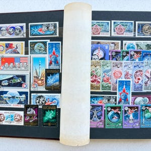 May include: A stamp album page featuring a collection of vintage postage stamps with space exploration themes, including depictions of astronauts, rockets, satellites, and celestial bodies, arranged on black pages with a central white divider.
