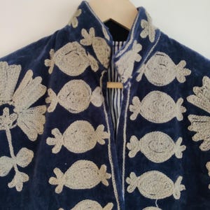 May include: A navy blue velvet jacket with intricate, light beige embroidered floral and candy-shaped designs. The jacket features a mandarin collar and a wooden toggle closure. The lining is a striped fabric. The jacket is hanging on a wooden hanger.