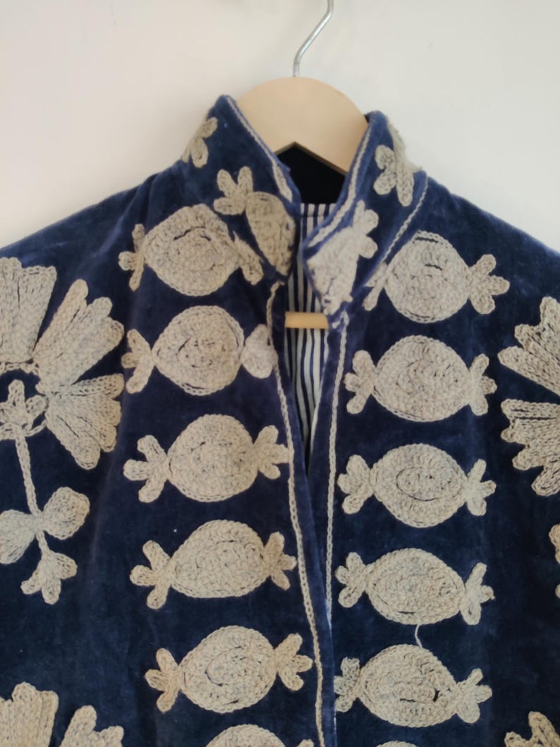 May include: A navy blue velvet jacket with intricate, light beige embroidered floral and candy-shaped designs. The jacket features a mandarin collar and a wooden toggle closure. The lining is a striped fabric. The jacket is hanging on a wooden hanger.
