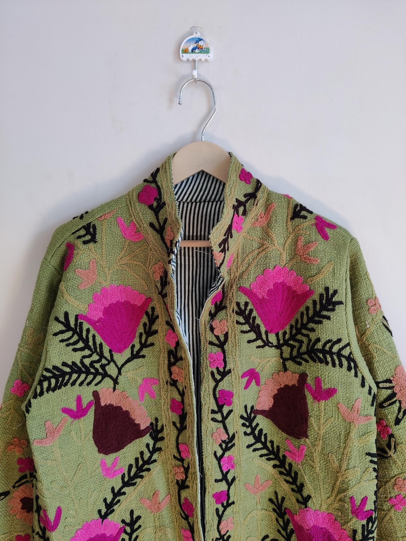 Puede incluir: Una chaqueta verde bordada con dise&ntilde;os florales rosas y marrones. La chaqueta tiene un cuello mandar&iacute;n y una cremallera.
