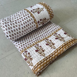 May include: A white and brown floral quilt, partially rolled, with a repeating pattern of small brown floral motifs. The quilt features a border with larger floral designs and a brown geometric pattern.