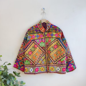 Puede incluir: Chaqueta bordada con un diseño geométrico y floral multicolor. Presenta un cuello mandarín y está adornada con bordados rosas, verdes, amarillos y azules. Colgada de una percha de madera, muestra su intrincado diseño y colores vibrantes.