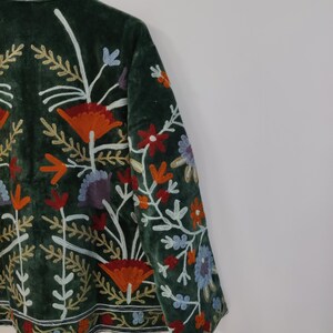 May include: Dark green velvet jacket with colorful floral embroidery. The jacket features orange, red, blue, and white embroidered flowers and leaves. The embroidery is detailed and covers the front and sleeves of the jacket.