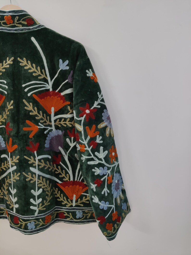 May include: Dark green velvet jacket with colorful floral embroidery. The jacket features orange, red, blue, and white embroidered flowers and leaves. The embroidery is detailed and covers the front and sleeves of the jacket.