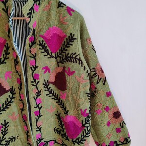 Puede incluir: Una chaqueta verde bordada con dise&ntilde;os florales rosas y marrones. La chaqueta tiene un cuello mandar&iacute;n y un forro a rayas blanco y negro.