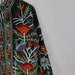 May include: Dark green velvet jacket with colorful floral embroidery. The jacket features a high collar and long sleeves. The embroidery includes orange, red, blue, and white flowers and leaves. The jacket is a unique piece of clothing.