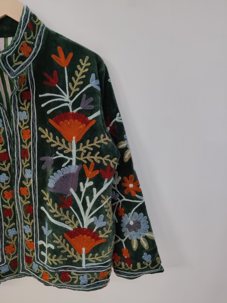 May include: Dark green velvet jacket with colorful floral embroidery. The jacket features a high collar and long sleeves. The embroidery includes orange, red, blue, and white flowers and leaves. The jacket is a unique piece of clothing.