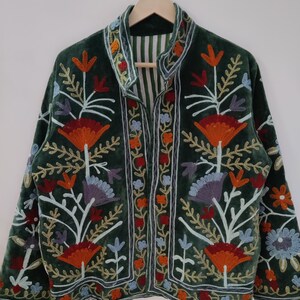 May include: A dark green velvet jacket with colorful floral embroidery. The jacket features a stand-up collar and a striped lining. The embroidery includes orange, red, blue, and purple flowers and leaves, creating a vibrant design.