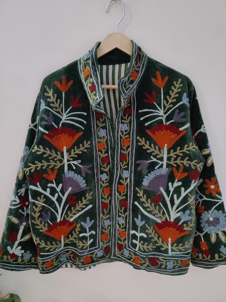 May include: A dark green velvet jacket with colorful floral embroidery. The jacket features a stand-up collar and a striped lining. The embroidery includes orange, red, blue, and purple flowers and leaves, creating a vibrant design.