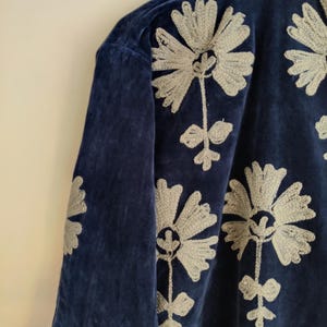 May include: A navy blue velvet jacket with embroidered floral designs in a light gray color. The jacket features a high collar and long sleeves. The floral embroidery is detailed and covers the front and sleeves of the jacket.