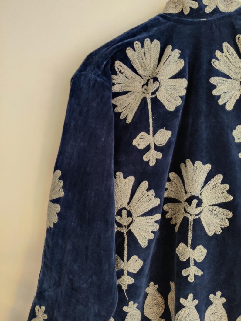 May include: A navy blue velvet jacket with embroidered floral designs in a light gray color. The jacket features a high collar and long sleeves. The floral embroidery is detailed and covers the front and sleeves of the jacket.