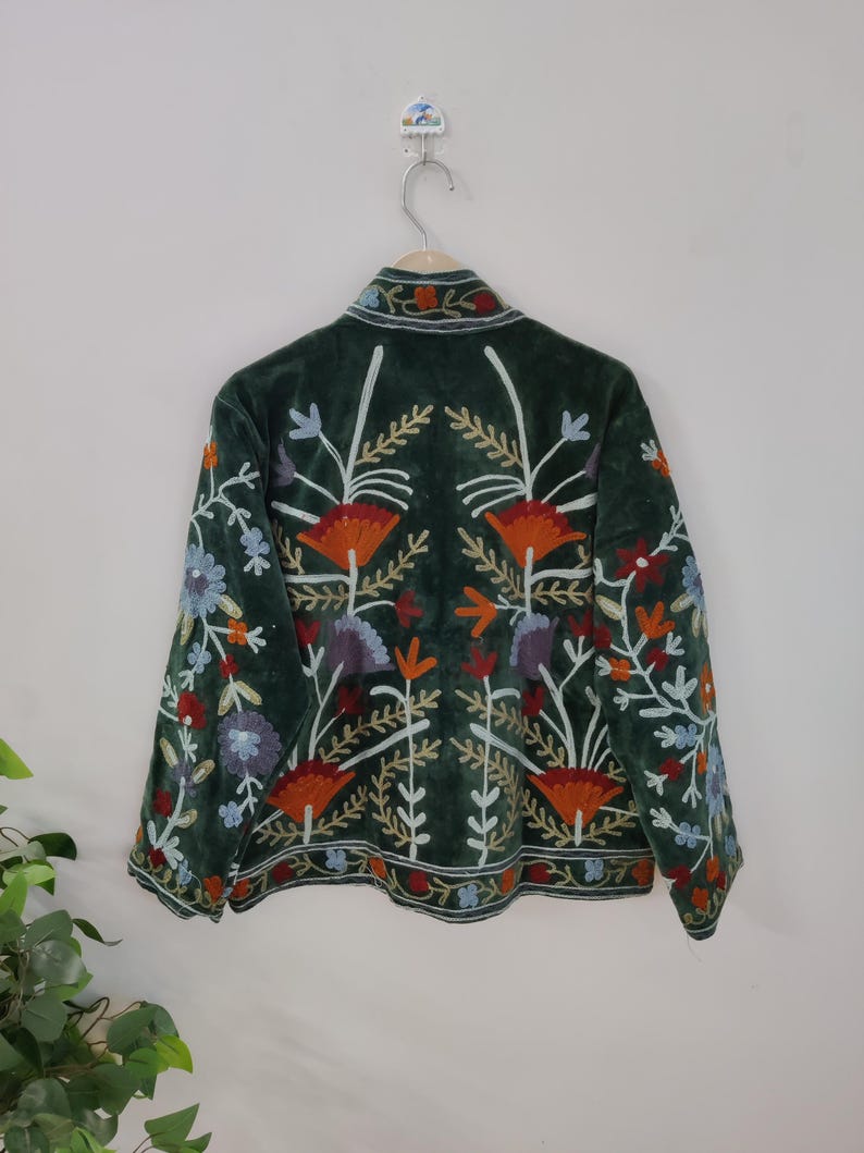 May include: A dark green jacket with colorful floral embroidery. The jacket features a velvet texture and a collar. The embroidery includes orange, blue, purple, and white flowers and leaves, creating a vibrant design.