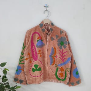 May include: A peach-coloured jacket with colourful floral embroidery. The jacket features a mandarin collar and a striped lining. The embroidery includes blue, green, pink, and orange floral designs. The jacket is hanging on a metal hanger.