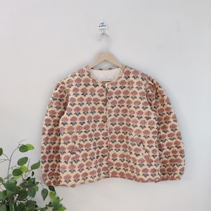 Könnte beinhalten: Beige Jacke mit floralem Muster, Rundhalsausschnitt und langen Ärmeln. Das Muster zeigt rote Blumen und grüne Blätter. Auf einem Holzbügel vor neutralem Hintergrund aufgehängt.