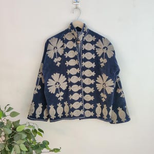 May include: A navy blue jacket with beige floral and candy-shaped embroidery. The jacket is hanging on a wooden hanger against a white wall. The embroidery is detailed and covers the front and sleeves of the jacket.