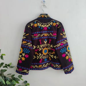 May include: A dark brown jacket with colorful floral embroidery. The embroidery features yellow, pink, blue, and orange flowers and leaf patterns. The jacket is hanging on a silver hanger against a white wall.