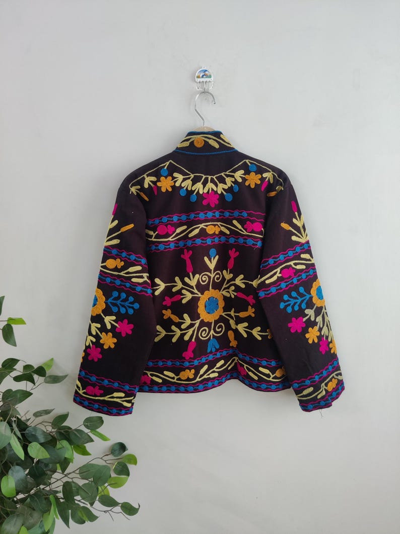 May include: A dark brown jacket with colorful floral embroidery. The embroidery features yellow, pink, blue, and orange flowers and leaf patterns. The jacket is hanging on a silver hanger against a white wall.