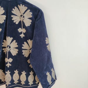 May include: A navy blue velvet jacket with intricate beige floral embroidery. The jacket features a high collar and long sleeves, with detailed floral patterns adorning the front, sleeves, and hemline. The embroidery adds a touch of elegance to the garment.