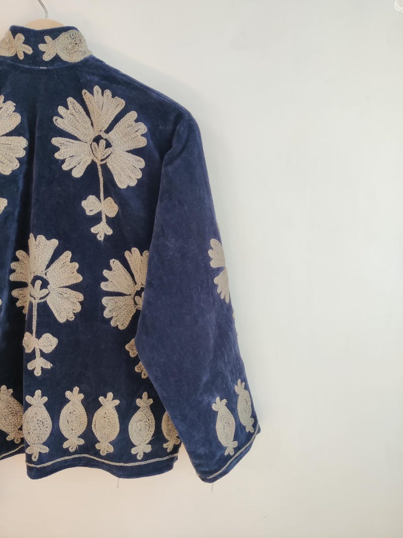 May include: A navy blue velvet jacket with intricate beige floral embroidery. The jacket features a high collar and long sleeves, with detailed floral patterns adorning the front, sleeves, and hemline. The embroidery adds a touch of elegance to the garment.