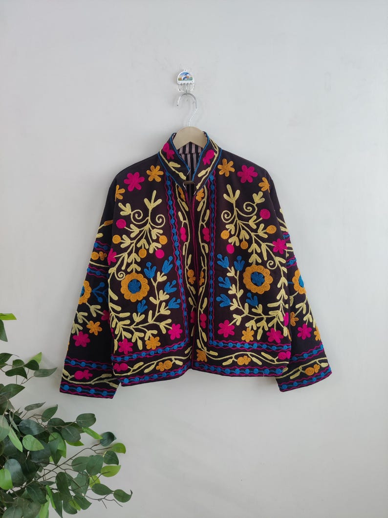 May include: A black jacket with colorful floral embroidery. The jacket features a high collar and is adorned with yellow, blue, and pink flowers and leaves. The embroidery is detailed and covers the front and sleeves of the jacket.