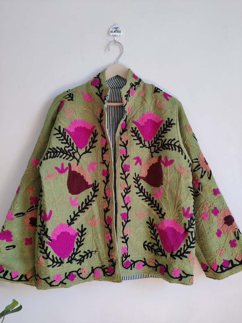 Puede incluir: Una chaqueta verde bordada con dise&ntilde;os florales rosas y marrones. La chaqueta tiene un cuello mandar&iacute;n y una cremallera frontal.