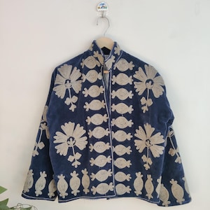 May include: A navy blue jacket with beige floral embroidery. The jacket features a high collar and is adorned with large flower and candy-shaped embroidered designs. The jacket is hanging on a wooden hanger against a white wall.