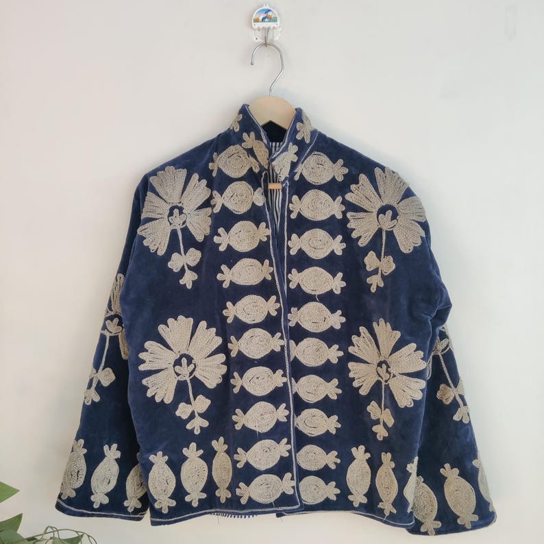 May include: A navy blue jacket with beige floral embroidery. The jacket features a high collar and is adorned with large flower and candy-shaped embroidered designs. The jacket is hanging on a wooden hanger against a white wall.