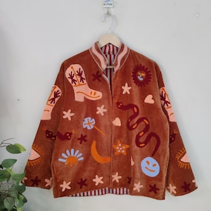 以下が含まれることがあります： カウボーイブーツ、ヘビ、太陽、星などの刺繍が施された茶色のベルベットジャケット。 ジャケットにはストライプの裏地とピンクのトリムが付いた襟が付いています。 ジャケットは木製のハンガーに吊るされています。