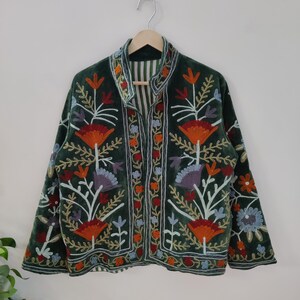 May include: A dark green velvet jacket with colorful floral embroidery. The jacket features orange, red, purple, and blue flower designs, with intricate detailing. The jacket is hanging on a wooden hanger against a white wall.