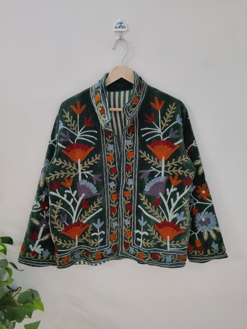 May include: A dark green velvet jacket with colorful floral embroidery. The jacket features orange, red, purple, and blue flower designs, with intricate detailing. The jacket is hanging on a wooden hanger against a white wall.