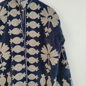 May include: A navy blue jacket with intricate beige embroidery. The embroidery features repeating candy-shaped and floral designs. The jacket is hanging on a wooden hanger, showcasing its detailed craftsmanship and unique style.