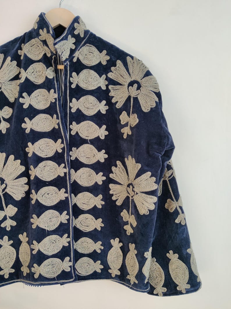 May include: A navy blue jacket with intricate beige embroidery. The embroidery features repeating candy-shaped and floral designs. The jacket is hanging on a wooden hanger, showcasing its detailed craftsmanship and unique style.