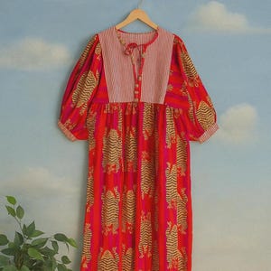 May include: A red dress with a tiger print design. The dress features a striped bodice with a tie closure and three-quarter sleeves. The dress is hanging on a wooden hanger against a blue sky backdrop.