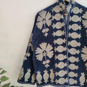 May include: Navy blue jacket with intricate silver embroidery. The jacket features floral and abstract patterns, with a high collar and long sleeves. The jacket is hanging on a wooden hanger against a white wall.