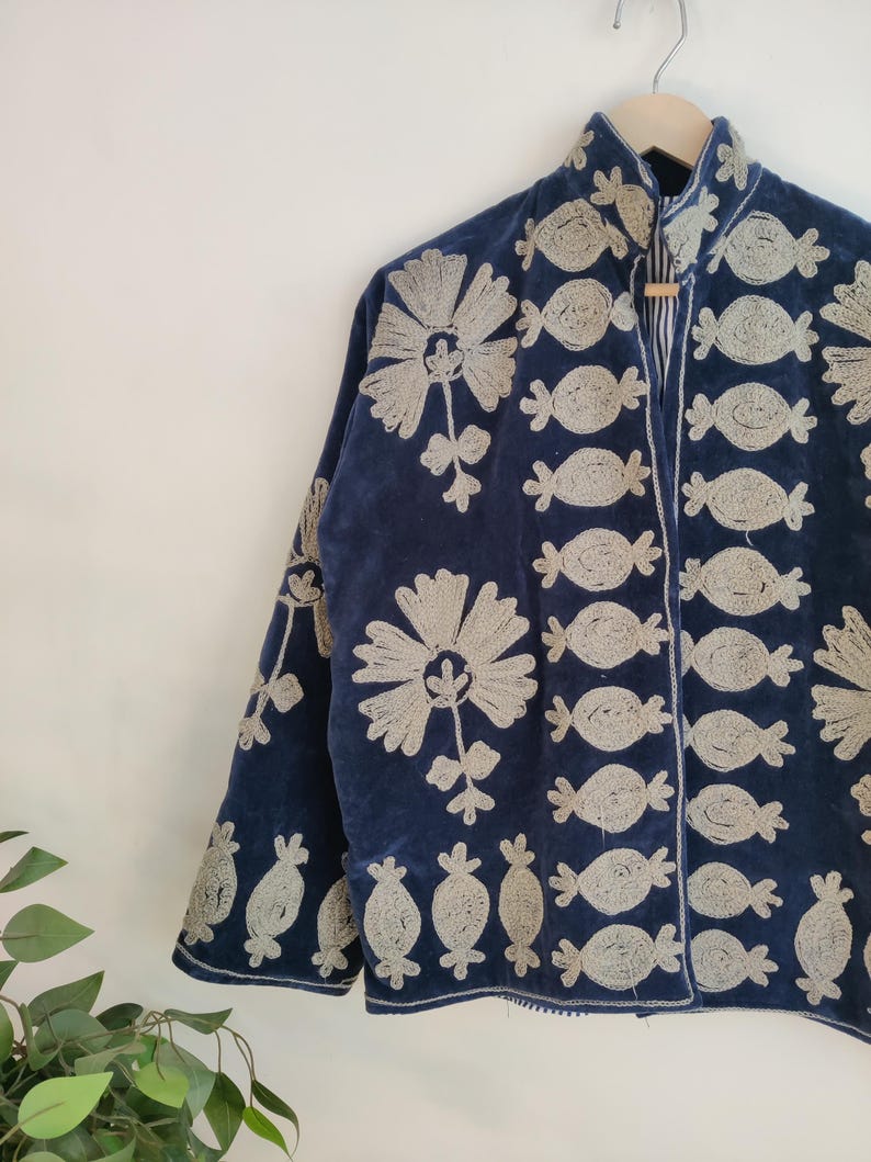 May include: Navy blue jacket with intricate silver embroidery. The jacket features floral and abstract patterns, with a high collar and long sleeves. The jacket is hanging on a wooden hanger against a white wall.