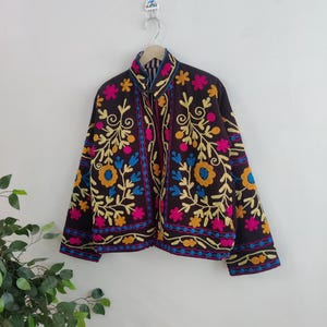 May include: A dark brown jacket with colorful floral embroidery. The jacket features yellow, pink, blue, and orange flowers and leaves. The jacket is hanging on a wooden hanger against a white wall.