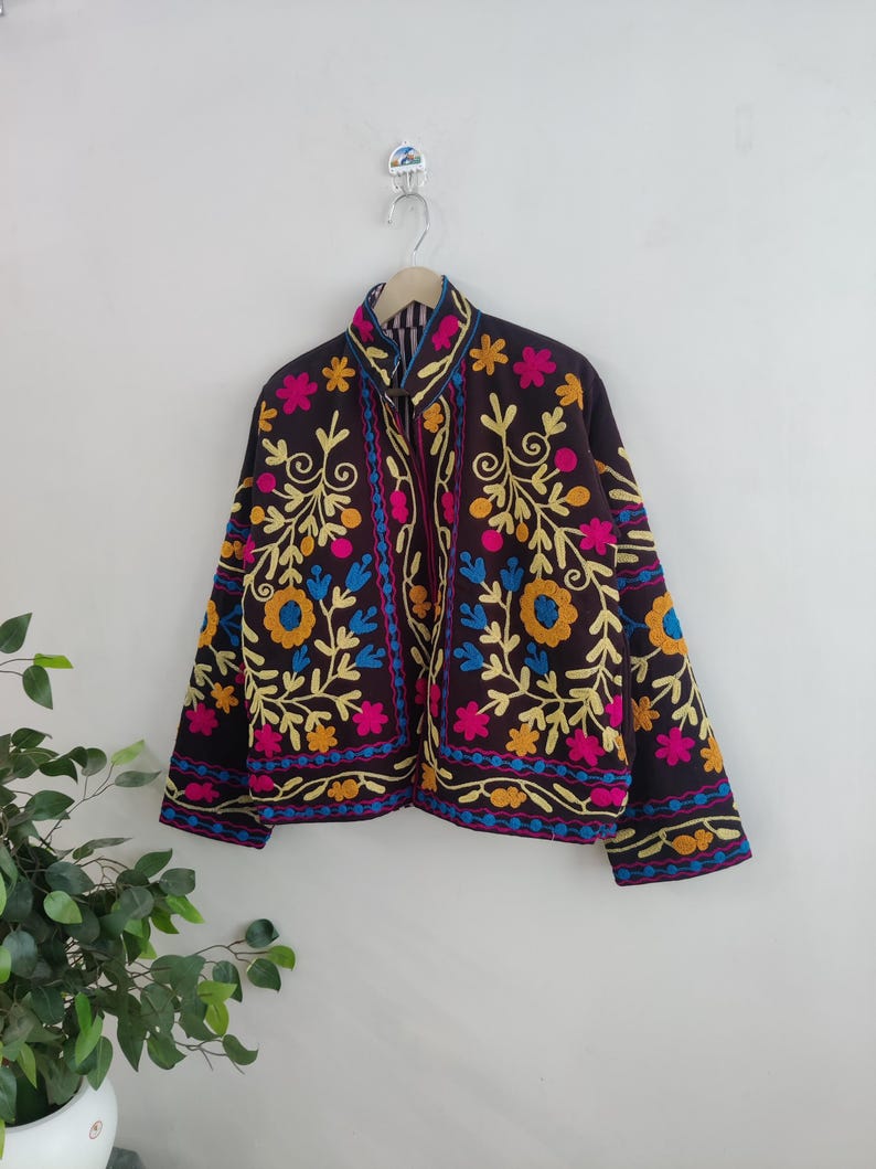 May include: A dark brown jacket with colorful floral embroidery. The jacket features yellow, pink, blue, and orange flowers and leaves. The jacket is hanging on a wooden hanger against a white wall.