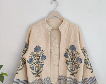 May include: A beige open-front jacket with blue floral patterns and blue striped trim. The jacket is hanging on a wooden hanger against a white background. The floral design is on the front and sleeves.