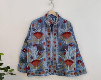 May include: A light blue velvet jacket with floral embroidery in red, orange, and gold. The jacket has a high collar and is hanging on a wooden hanger. The embroidery features stylized flowers and leaves.